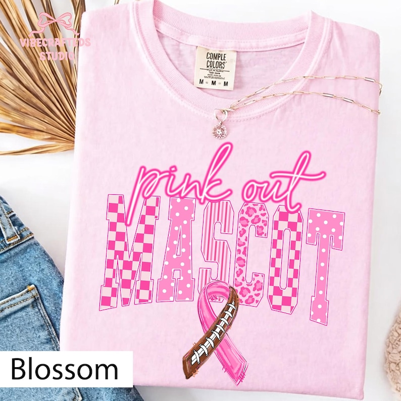 May include: A light pink short-sleeved t-shirt featuring the text pink out MASCOT in various pink patterns including checkerboard, stripes, leopard print, and polka dots. A pink awareness ribbon designed with football laces is centered below the text.