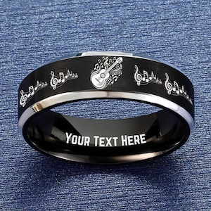 May include: Black and silver ring with musical notes and a guitar design. The ring has the text "YOUR TEXT HERE" engraved on the inside. The ring is on a blue textured surface.