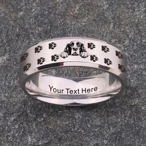 Puede incluir: Un anillo plateado con un diseño de perro y huellas de patas. El anillo presenta una ilustración detallada de la cara de un perro y huellas de patas alrededor de la banda. El texto "Your Text Here" está grabado en el interior.