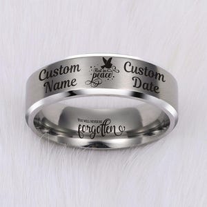 May include: A silver-colored ring with engraved text and a dove symbol. The ring features the words "Custom Name" and "Custom Date" on the outer band, with "Rest in Peace" and a dove symbol. The inner band reads "You will never be forgotten."