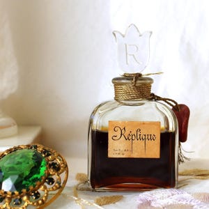 May include: A vintage perfume bottle with a clear glass stopper and a label that reads "Réplique." The bottle is filled with a dark liquid and is adorned with a decorative ring featuring a large green stone and gold accents.