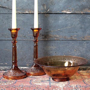 May include: Two amber glass candlesticks with tall white taper candles and a matching amber glass bowl with a decorative rim. The items feature a translucent finish and intricate embossed patterns on the bases and the bowl's edge.