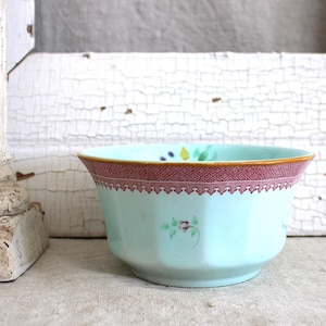May include: A light blue ceramic bowl featuring a wide, intricate reddish-pink patterned border along the upper rim and a small, delicate floral motif painted in green and pink on the exterior side of the bowl, which has a faceted shape.