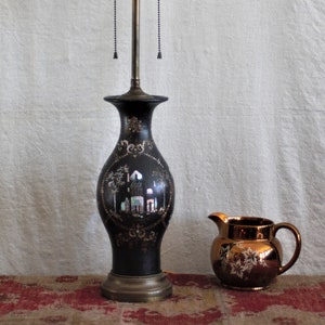 May include: A black table lamp with a decorative design and a brass base. The lamp has a three-bulb fixture and a matching decorative pitcher with a floral design. The lamp and pitcher are on a patterned rug.