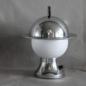Peut inclure: Une lampe de table composée d'un dôme supérieur en chrome poli avec un rebord circulaire plat et un petit embout noir. La partie centrale comprend un abat-jour sphérique en verre dépoli blanc, soutenu par une base évasée en chrome réfléchissant.