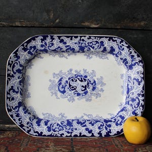 May include: A rectangular ceramic serving platter with rounded corners, featuring a white base and a deep blue intricate floral scrollwork pattern along the border and in the center. A single yellow apple is positioned next to the platter.