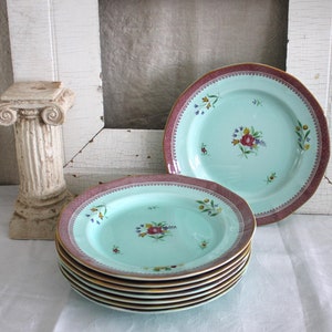 May include: A stack of six aqua-colored plates with a floral design and a burgundy rim. Two additional plates are visible, one stacked on top and another to the side. A small decorative column and a white distressed frame are in the background.