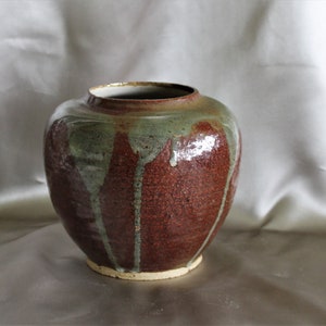 Könnte beinhalten: Eine braune Keramikvase mit grüner Glasur und weißen Glasurtröpfchen. Die Vase hat einen runden Körper und eine breite Öffnung.