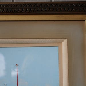 Puede incluir: Primer plano de un marco de imagen de color dorado con un borde decorativo. El marco rodea una pintura de un cielo azul con un poste rojo y una nube blanca.