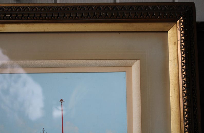Puede incluir: Primer plano de un marco de imagen de color dorado con un borde decorativo. El marco rodea una pintura de un cielo azul con un poste rojo y una nube blanca.
