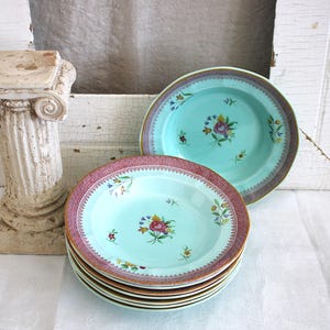 May include: A stack of aqua-colored ceramic plates with a floral design and a decorative border. The plates are stacked on a white surface, with a decorative column in the background.