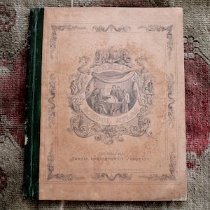 Könnte beinhalten: Antiker Schulatlas mit grünem Rücken und beigefarbenem Einband. Der Einband zeigt eine verzierte Illustration mit den Worten "School Atlas" und "Philadelphia" aufgedruckt. Das Buch liegt auf einem gemusterten Teppich.