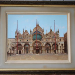 Puede incluir: Una pintura enmarcada que representa un gran edificio adornado con m&uacute;ltiples c&uacute;pulas y torres. El edificio est&aacute; pintado en tonos beige y marr&oacute;n, con banderas rojas ondeando desde la cima de las torres. El edificio se encuentra sobre un fondo de cielo azul con nubes blancas.