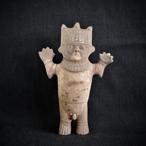 Pode incluir: Figura desgastada, de aspeto antigo, de uma figura em pé com os braços levantados. A figura tem uma coroa, um colar de contas e uma barba. A cor é uma mistura de castanho, cinzento e bege, com desgaste visível.