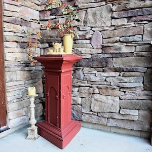 May include: A tall, red, rectangular wooden pedestal with arched panel detailing. On top sits a yellow vase with orange berry branches. Beside the pedestal stands a distressed white candle holder with a cream-colored pillar candle on top.