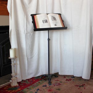 May include: A black metal music stand holds an open book with illustrations. The stand has a tall, adjustable pole and a decorative base. A white candle sits on a decorative stand to the left. The background includes a white curtain and a patterned rug.