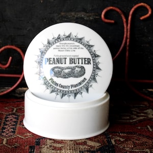 Może przedstawiać: Biały ceramiczny słoik z masłem orzechowym z pokrywką z napisem "PEANUT BUTTER" i ilustracją orzeszków ziemnych. Słoik jest cylindryczny z zaokrągloną górą.