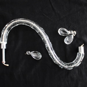 May include: A crystal chandelier arm with a clear glass design and three crystal drops. The arm is curved and has a silver metal connector at the end.