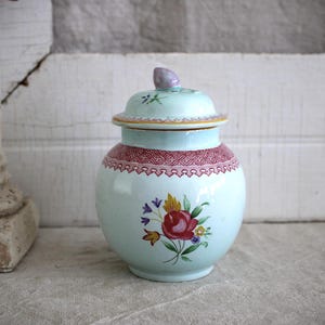May include: A light blue ceramic jar with a lid, featuring a red patterned border around the neck and a hand-painted floral motif on the front. The lid has a small, light purple finial at the top, and the body has a rounded, bulbous shape.