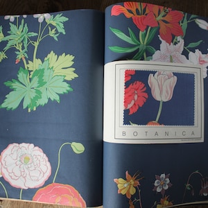 May include: An open book displaying pages with a dark blue background featuring various botanical illustrations of flowers and leaves in shades of pink, yellow, and green. A rectangular paper insert with a tulip graphic and the text BOTANICA.