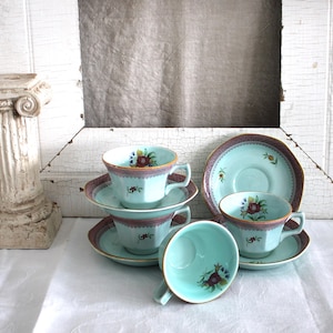 May include: A collection of vintage turquoise teacups and saucers. Each cup and saucer features a floral design with a burgundy and gold trim. The teacups are arranged on a white surface, with a decorative column in the background.