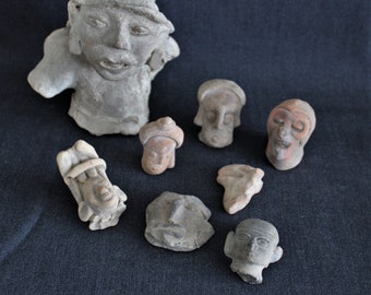 Puede incluir: Colección de siete pequeñas esculturas de terracota de cabezas humanas con diversas expresiones faciales. Las esculturas están todas hechas a mano y tienen un aspecto rústico y terroso.