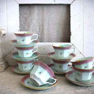 May include: A set of eight vintage teacups and saucers with floral patterns in shades of pink, green, and white. The cups and saucers are stacked in a pyramid shape on a light blue surface.