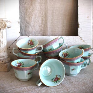 May include: A collection of light blue ceramic teacups featuring a decorative reddish-brown patterned border along the rim and small floral motifs painted on the exterior and interior surfaces. Each cup has a handle and a faceted, octagonal base.