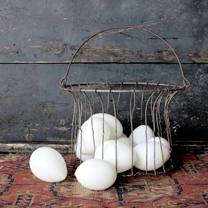 Può includere: Un cestino vintage per uova in filo metallico pieno di uova bianche. Il cestino ha un manico e poggia su un tappeto fantasia. Lo sfondo è una parete di legno scuro e usurato.