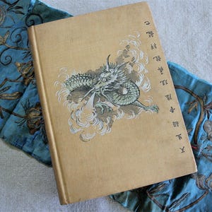 May include: A tan hardcover book featuring a green dragon illustration surrounded by white clouds and vertical Asian characters on the cover, resting on a piece of blue fabric with gold embroidered floral and vine patterns.