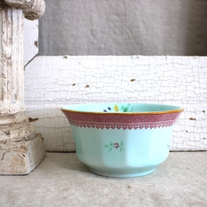 May include: A light blue porcelain bowl with a pink and gold rim and floral designs. The bowl is sitting on a white surface.