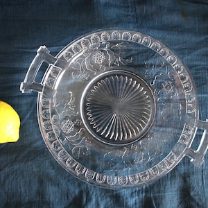 Pode incluir: Uma bandeja de servir de vidro transparente com um padrão floral decorativo e duas alças. A bandeja tem uma seção central elevada com um design de raios de sol.