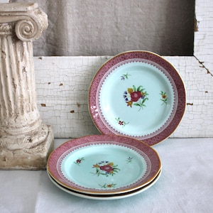 May include: Three antique porcelain plates with a light blue center and a decorative burgundy border. Each plate features a floral design in the center. The plates have a gold rim and are stacked on top of each other.