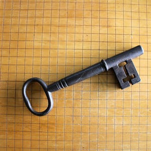 May include: A dark grey metal skeleton key with an oval bow and a complex bit, positioned next to a wooden ruler showing a length of approximately 16.5 cm from the tip of the bit to the end of the oval handle on the grid surface.