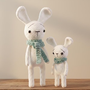 May include: Two white crochet bunny dolls. The larger bunny wears a light green scarf, and the smaller bunny also wears a light green scarf. Both bunnies have black eyes and a black stitched nose and mouth.