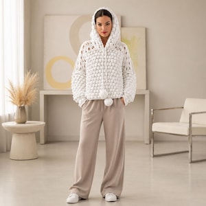 Könnte beinhalten: Eine weiße Kapuzenjacke aus Häkelarbeit mit einem strukturierten Noppenmuster. Die Jacke hat lange Ärmel und einen Reißverschluss vorne. Das Model trägt eine beige Hose mit weitem Bein und weiße Turnschuhe. Die Jacke hat einen Bommel am unteren Saum.