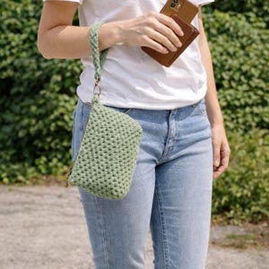 Può includere: Una pochette all'uncinetto verde chiaro con un cinturino da polso intrecciato abbinato. La pochette ha una chiusura a cerniera e un anello color oro che collega il cinturino. La persona indossa jeans blu e una maglietta bianca.