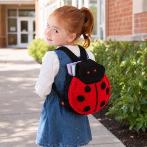 May include: A red and black crochet ladybug backpack with black straps and a black top. The backpack has large black spots and is worn by a child. The child is wearing a denim dress and white shirt.