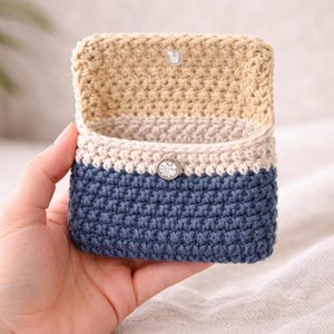 Puede incluir: Una pequeña caja de ganchillo abierta en tonos azules y beige. La caja se mantiene abierta, revelando el interior. La solapa superior se asegura con un botón transparente. Las puntadas de ganchillo son visibles, creando una superficie texturizada. La caja es probablemente hecha a mano.