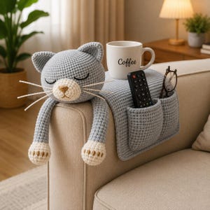 May include: A gray crocheted cat-shaped sofa armrest organizer with a cream-colored muzzle, long arms, and white-tipped paws. The organizer features two pockets containing a black remote control and a pair of glasses, with a white mug labeled Coffee.