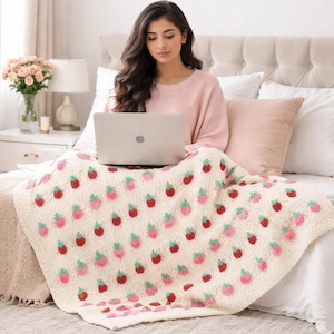 Op de afbeelding: Een crèmekleurige, gehaakte deken met een herhalend patroon van rode en roze aardbeien met groene bladeren. De deken is over een bed gedrapeerd en een persoon gebruikt een laptop terwijl hij op het bed zit.