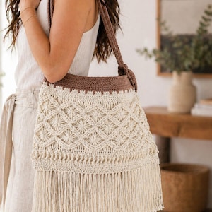 Peut inclure: Un sac en crochet crème avec un dessus et une bandoulière marron. Le sac présente un motif géométrique et une frange en bas. La bandoulière est nouée en haut. Le sac est un sac à bandoulière.