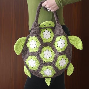May include: A handmade crochet turtle-shaped bag in shades of brown, green, and white. The bag features a turtle head and flipper-like appendages. The body is made of hexagonal panels with white flower-like designs.