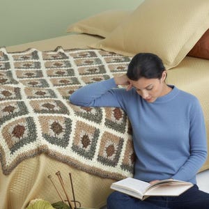 May include: A handmade crochet blanket with a geometric pattern in shades of brown, green, and cream rests on a bed. A green container holds various colored yarn balls and knitting needles. The scene suggests a cozy, handcrafted theme.