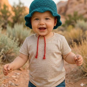 Puede incluir: Un gorro de bebé de ganchillo color turquesa con un borde festoneado y lazos rojos. El bebé lleva una camiseta beige y vaqueros azules. El bebé sonríe y mira a la cámara.