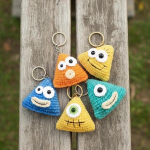 May include: A collection of five handmade, crocheted triangle keychains with cartoon faces. The keychains are in various colors: blue, orange, yellow, and teal. Each keychain has a metal ring for attaching keys.