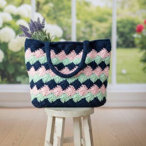 May include: A navy blue, pink, and mint green crocheted tote bag with a geometric pattern. The bag has a matching navy blue handle and is filled with lavender sprigs. The bag is sitting on a small, white wooden stool.