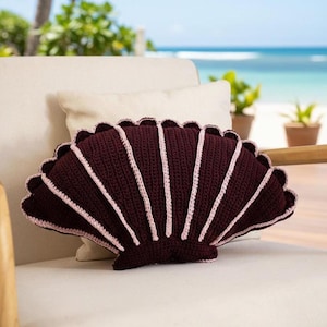 May include: A burgundy crochet seashell pillow with light pink accents. The pillow is fan-shaped with a scalloped edge and is resting on a white chair. The background shows a beach scene with the ocean and plants.