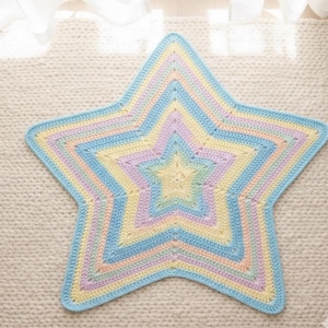 May include: A star-shaped crochet blanket in pastel shades. The blanket has layers of soft colours, including yellow, blue, pink, and lavender. The edges are light blue. The blanket is likely handmade and suitable for a baby or child.
