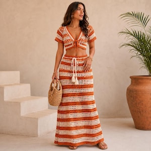 May include: A two-piece crochet set consisting of a short-sleeved, V-neck cropped top with a front tie and a high-waisted, floor-length maxi skirt. Both pieces feature horizontal stripes in burnt orange and cream with an open-knit texture.